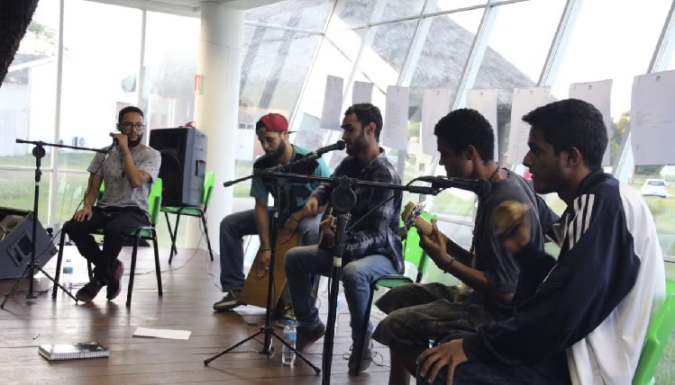 Cinco estudantes homens estão sentados tocando instrumentos de percussão e cantando no microfone.