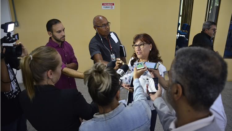 No centro da foto, a reitora da FURG, Cleuza Maria Sobral Dias, está rodeada por repórteres que carregam microfones e smartphones. À esquerda, um cameraman aponta a câmera para a reitora.