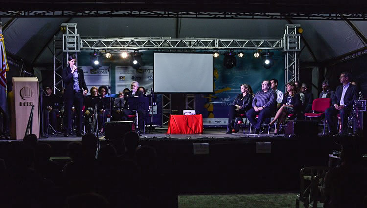 A foto é escura, uma foto feita à noite. Em uma palco iluminado, há diversas pessoas sentadas. Um homem fala ao microfone. 