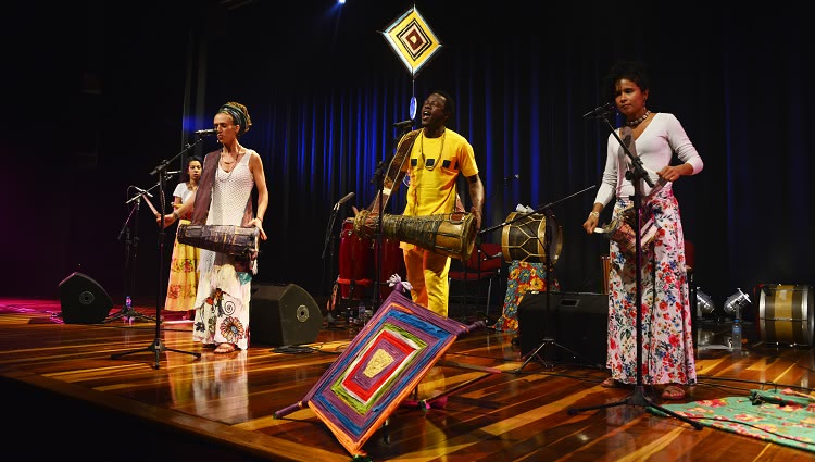 Em cima do palco, as quatro mulheres do grupo Três Marias tocam seus instrumentos de percussão em um palco decorado com elementos coloridos, enquanto o nigeriano Akin canta e toca.