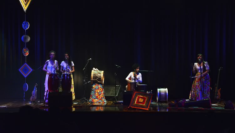 Foto do palco onde o grupo Três Marias. Duas cantoras em pé, seguidas por uma sentada e outra em pé, todas com seus instrumentos.