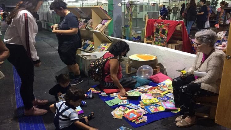 Crianças, uma senhora e outros visitantes em frente ao estande do Troca de Livros na 46ª Feira do Livro da FURG.