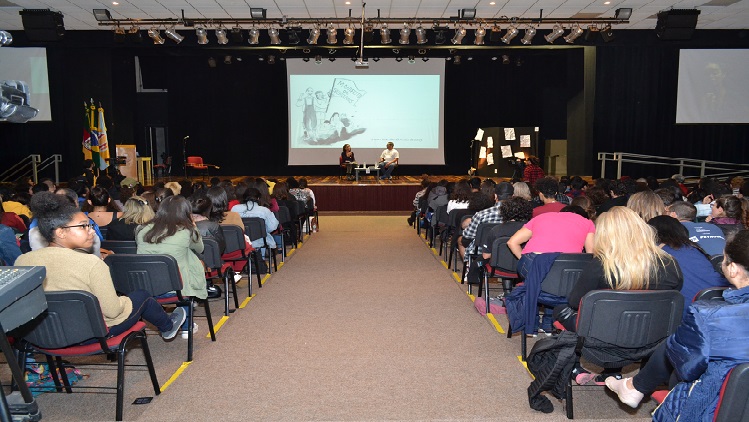 A foto mostra em primeiro plano pessoas sentadas olhando em direção ao palco onde está Sergio Vaz e a professora Luciana Coronel.
