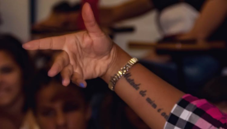 A foto mostra em destaque a mão de uma mulher gesticulando enquanto fala. Ela usa um relógio e tem uma tatuagem no antebraço.