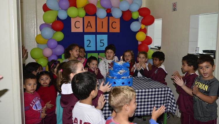 A foto mostra crianças na volta do bolo de aniversário no refeitório do Caic.