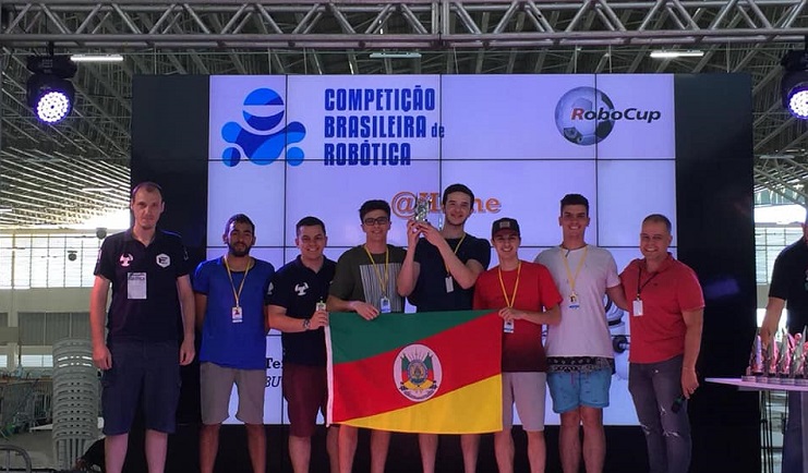 A foto mostra alunos integrantes do Nautec recebendo um prêmio por um trabalho apresentado. O grupo segura o troféu e também a bandeira do Rio Grande do Sul.