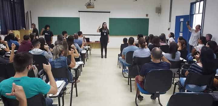 A foto mostra uma sala de aula com a intérprete em pé, no centro da sala, ensinando a linguagem dos sinais para os alunos que estão sentados a sua frente.